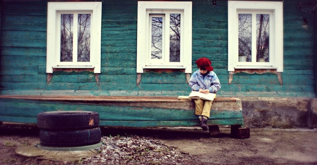 Child reading a book outside a rustic green house, under a street sign, conveying solitude and curiosity.