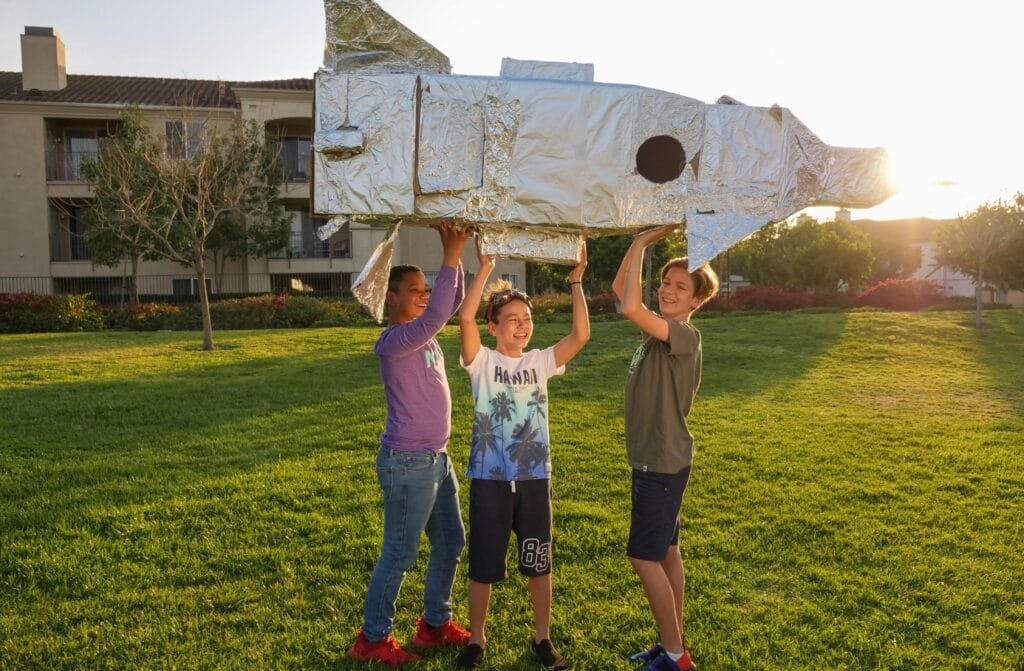 Three boys joyfully lift a homemade cardboard rocket while playing outdoors in a sunny park.