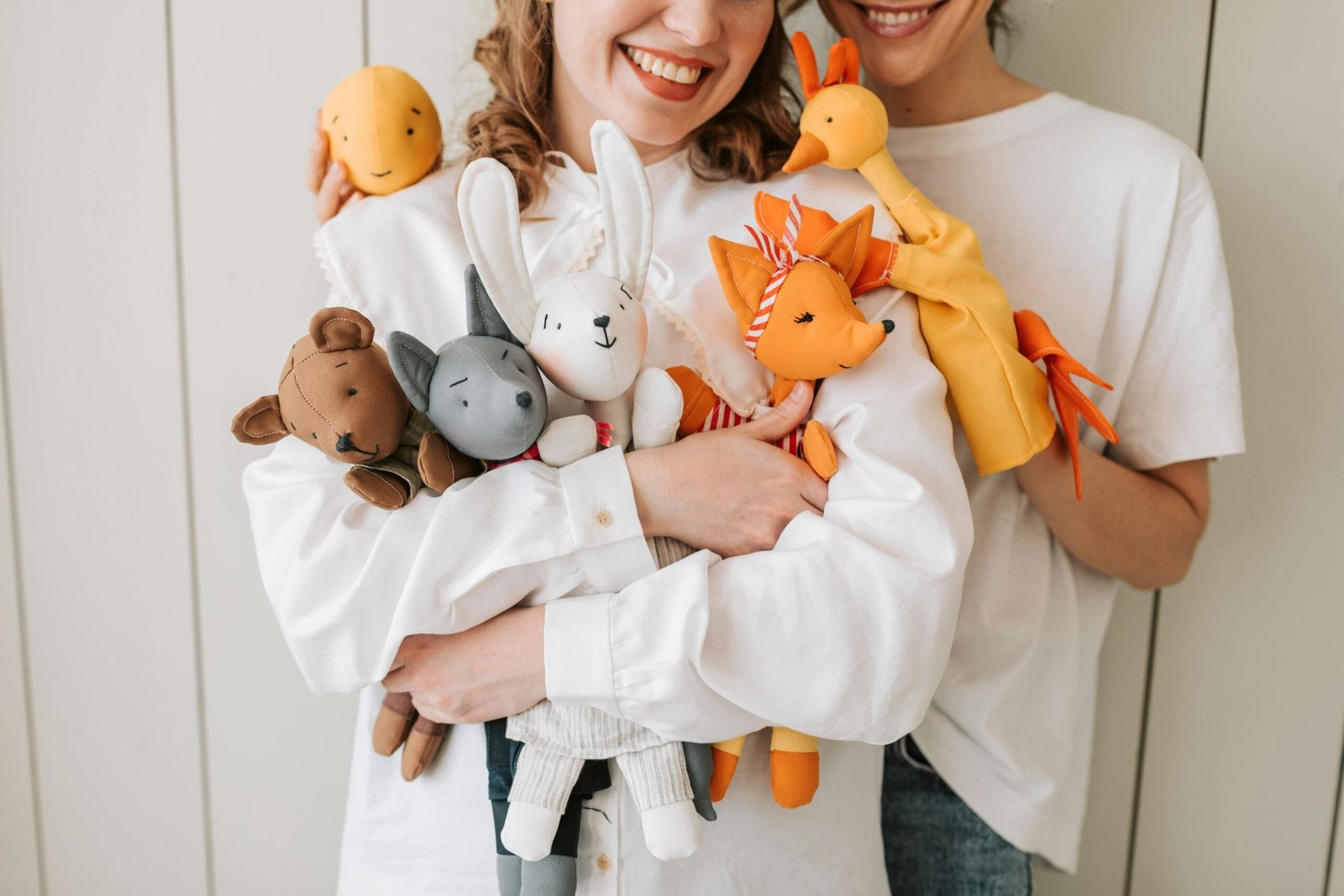 Two women smiling and holding a variety of colorful plush and hand puppets indoors.